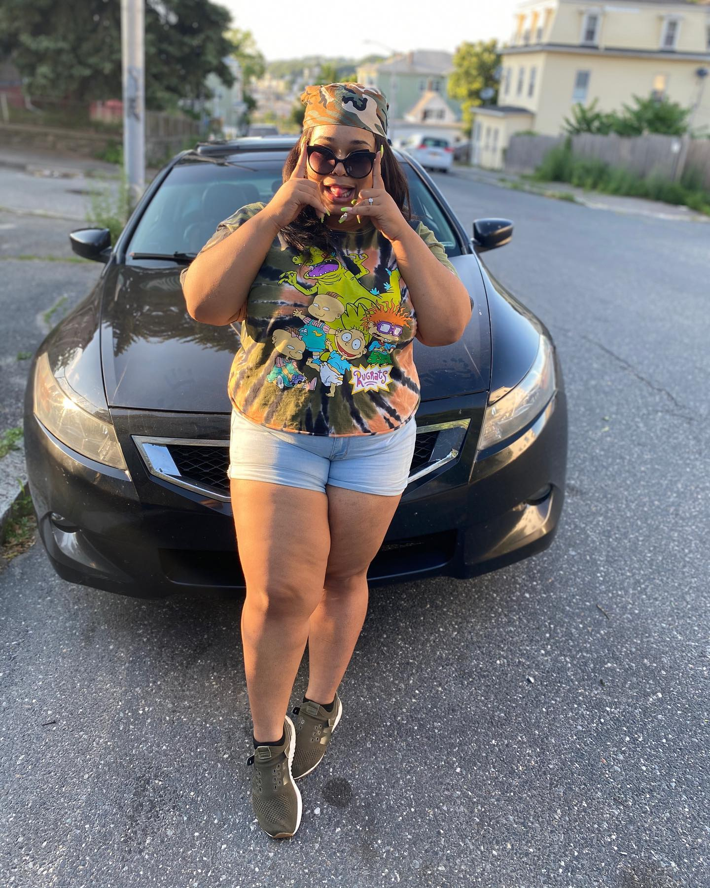 Woman in Rugrats tee, shorts, and bandana posing playfully by a car.