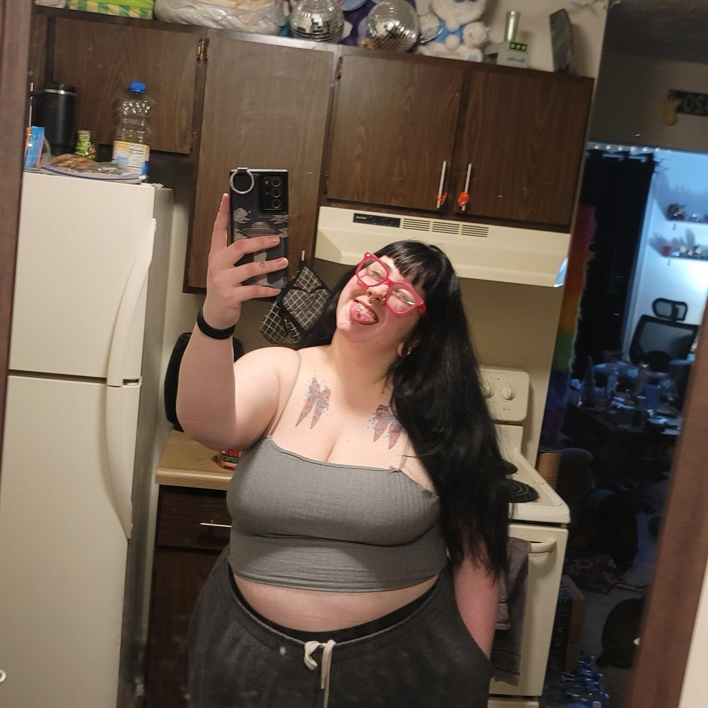 Woman in grey crop top and pink glasses taking a mirror selfie.