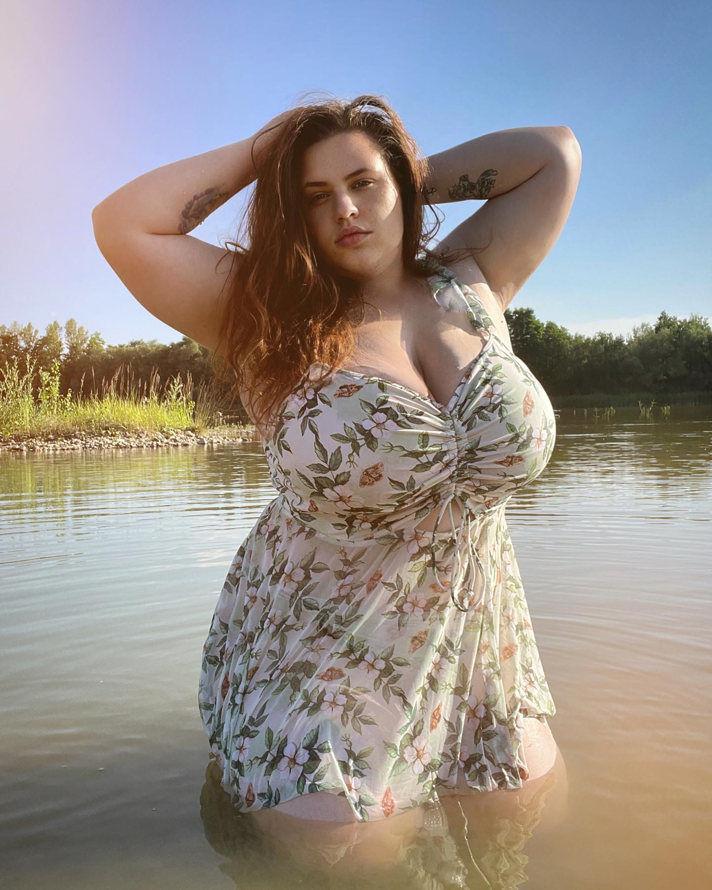 Woman in a floral dress standing in water with hands behind head.